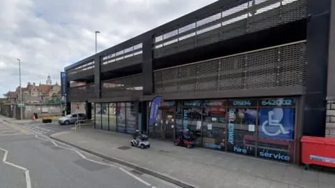Grey brick built shops with a car park on top.  mobility scooter can be seen on the pavement.