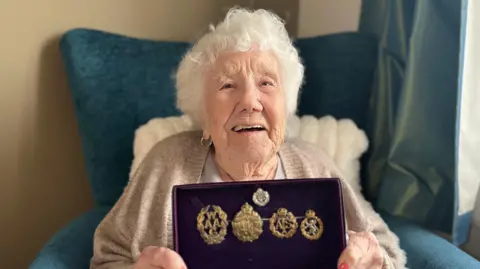 Elsie Wilkins is sitting on a turquoise armchair and is holding a rectangle box which has four gold medals in it. 