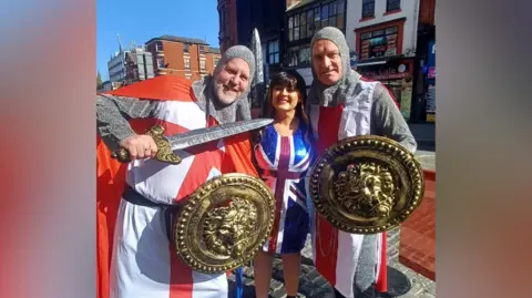 Lisa Hughes Three people in fancy dress. Two men are dressed as knights with St George's flags and swords. Lisa wears a brown wig and a Union Jack dress.