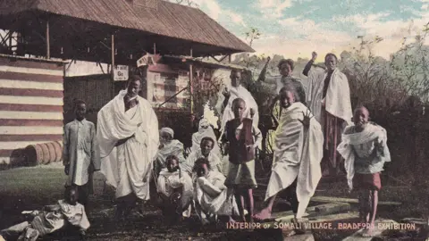 A group of people in traditional African dress, mostly made of white sheets of cloth wrapped around them. There are men, women and children among them. They are posing in front of a wooden hut. The black and white photo is old and faded. In the corner it is marked with the words 'Interior of Somali Village, Bradford Exhibition'