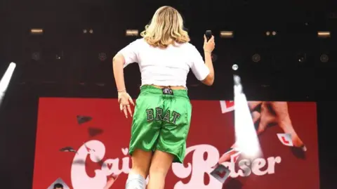 Getty Images Singer Caity Baser seen from behind on stage, in green silk shorts with the word brat sewn onto them, plus a white T shirt