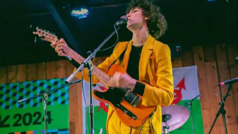 Otto Aday stands in front of a Welsh flag on stage at Texas' South by Southwest festival SXSW. He is wearing a yellow suit, playing an orange guitar, and singing in to a microphone.