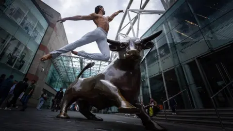 A male dancer wearing white trousers leaps high into the air next to a bull statue with glass buildings behind him