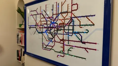 A framed map of the London transport network, with dozens of multicoloured LED lights tracking the live locations of individual services. 