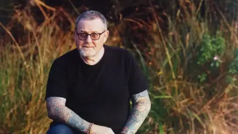 East Suffolk and North Essex NHS Foundation Trust David Spaul has short grey hair and black glasses. He has tattooed arms and is wearing a black T-shirt. He is crouching outside with long grass behind him.