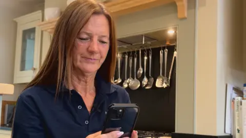 Maggie Pavlou is in a kitchen looking down at a phone. She has light brown hair and is wearing a navy blue polo shirt.