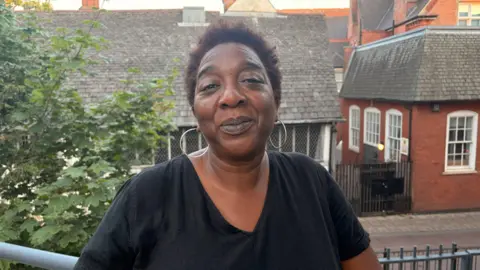 Lisa is stood on a balcony with the Guildhall in Leicester behind her. She is wearing hoop earrings and a black t-shirt. 