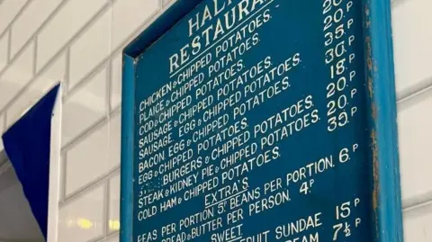 Blue wooden framed board hangs on a white tiled wall with the prices of items on sale at a fish and chip shop