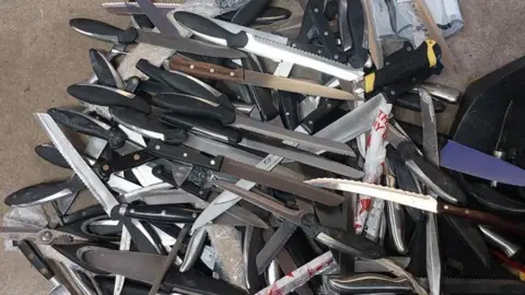 A large pile of weapons, mainly large kitchen knives with a mix of smooth and serrated edges. 