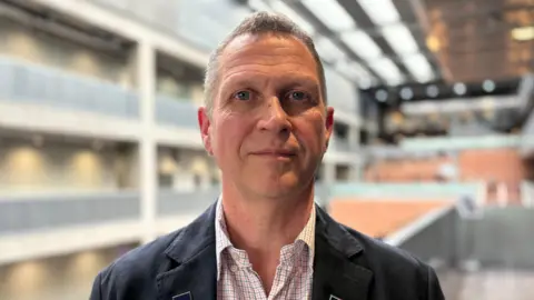 Close-up of Mike Corbett standing indoors in a modern, multi-level building, wearing a dark jacket and patterned shirt, with a blurred architectural background