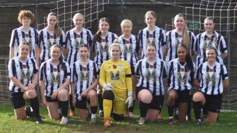 Raymond Cunningham A team photo. The front row are on bended knee, the back row are standing. The women wear black and white striped tops, apart from the goalkeeper in the centre who wears yellow