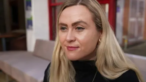 Rhi has long, blonde hair and is wearing a black roll-neck top. She is stood in a room with red-framed windows behind her, which are out of focus.