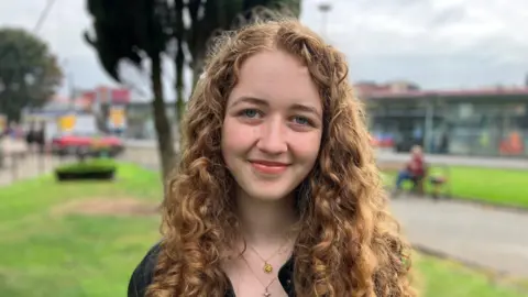 Amelia, who has long curly brown hair and is wearing a black top with necklaces, smiles while stood outdoors in a public park. 