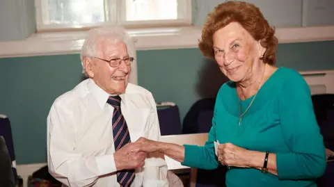 The Croft Mr Bickley, in white shirt and striped tie, holds a woman's hand. She has auburn hair and a green top with a thin gold necklace and a watch on her wrist. She slightly smiles at the camera while Mr Bickley looks at her