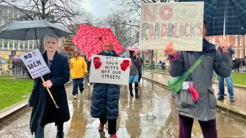 Three women are in the front of a march holding home-made signs reading "No to roadblocks" and "Keep your mitts off our streets". They are also carrying umbrellas.
