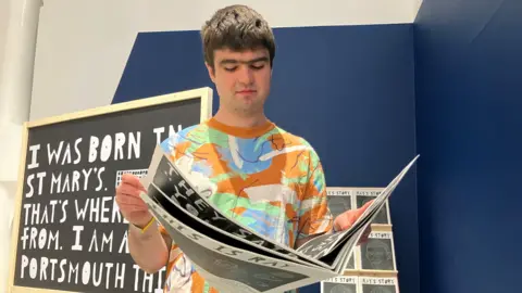 Orlando Richardson is wearing a bright colourful t-shirt and is holding his artwork of a newspaper inspired by a person called Ray.