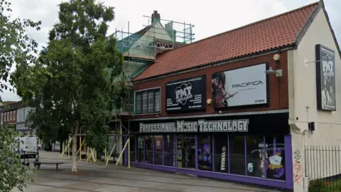 The outside of the Professional Music Technology store in Norwich. It has a large sign with the stores title, and purple borders around the window. There is a tree to the left of it. 
