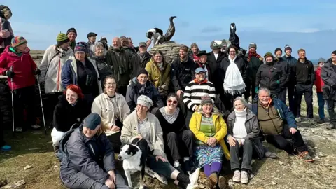 Rob Flett/BBC A mixed group of approximately forty people in outdoor clothes are gathered around the Great Auk statue and cairn. Some are sitting on the rocky ground and all are looking at the camera.