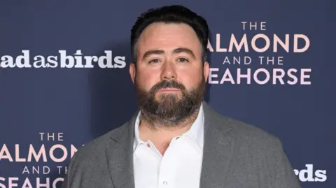 Getty Images Celyn Jones wearing a grey blazer and a white shirt. He is standing in front of a dark blue screen. He has short dark hair and a short beard.