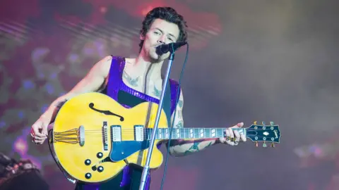 PA Media Harry Styles on stage singing into a microphone and holding a bass guitar, wearing a black and purple set of overalls 