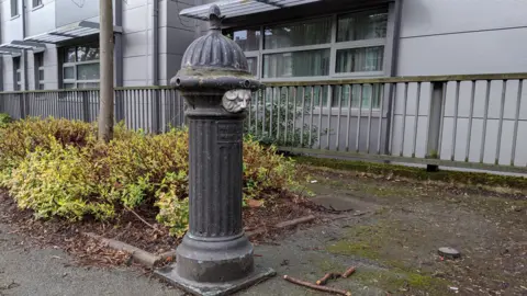 Ben Ellwood A black metal cylinder standing upright on a paved street in front of metal railings and a small bush, with a grey building behind it