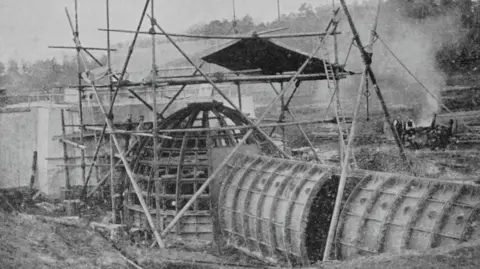 The Builder's Journal A historic black-and-white photograph showing the underwater room under construction. Scaffolding surrounds the domed structure while the tunnel leading to the room extends across the foreground. Construction work is taking place around the site before the lake was created.
