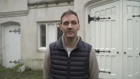 A man with greying hair and wearing a gillet over a long-sleeved shirt. He is standing outside a property.