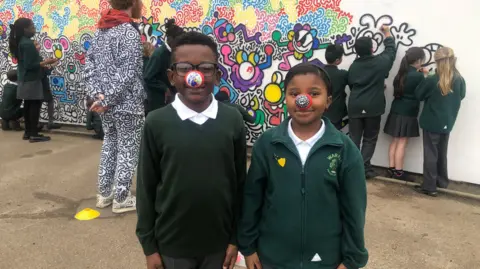 Two schoolchildren can be seen facing the camera, wearing comic relief noses that have had their own colours and designs drawn on. Schoolchildren can be seen in the background drawing on the mural with Mr Doodle, in his black and white suit, watching on.