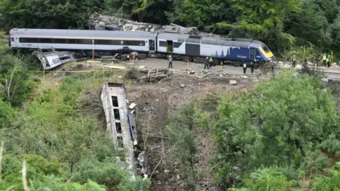 PA Media Stonehaven derailment - image of a train on tracks in woodland, and also a burned carriage down a slope.
