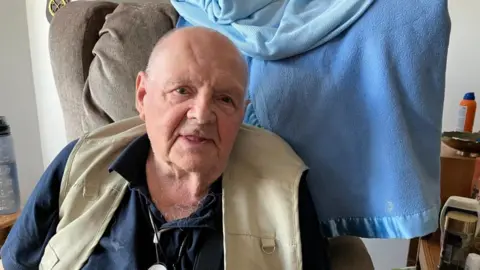 A man sitting in a grey armchair. He has a beige vest jacket on over a navy blue shirt.