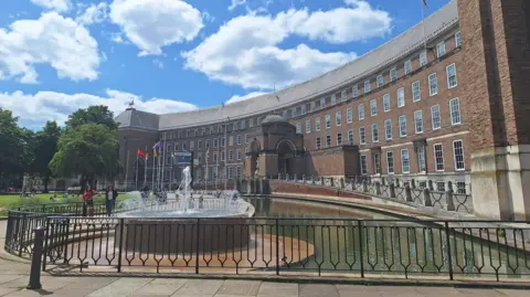 Bristol City Hall, which is a large curved brick building with lots of windows and a water feature and fountain in front of it. It is a sunny day with blue skies and fluffy white clouds. There are some flags flying on flagpoles outside, and a large lawn area with trees.