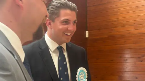 George Finch wears a Reform rosette and a navy tie with the Warwickshire bear and staff. He wars a dark suit jacket and a white shirt and has brown slicked back hair. He is pictures at the count for the local elections.