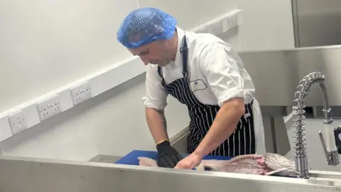 Grimsby Institute A man in an apron and a hair net, wearing a white chef's outfit, prepares a fish for cooking in a stainless steel kitchen area. 