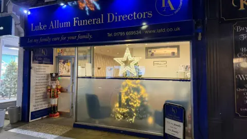 Luke Allum A blue shop front. It is a funeral home. In the doorway is a nutcracker statue. 