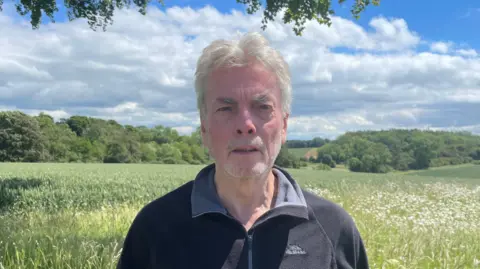 BBC/Amelia Riley A man with grey hair, dressed in a black zip-up fleece, stands in a green field with trees and rolling countryside behind under a partly cloudy blue sky.