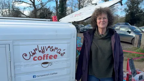 Hannah Roe/BBC Jane Killick pictured beside her cart, 'Curlyveras Coffee'. The cart is white and has the name emblazoned on the side. Jane has dark, curly hair and is wearing a purple coat and green jumper.