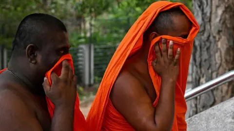 Two monks, wearing traditional orange attire, try to conceal their faces as they walk up stone stairway outside a building which can't be seen