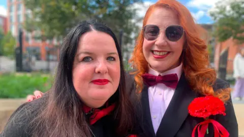 A woman with dark hair and a woman with red hair - both wear red bow ties and carnations and red lipstick