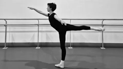 A boy is performing a ballet pose in a dance studio. He is standing on one leg with the other leg extended straight behind them at hip height, forming a horizontal line. One arm is stretched forward while the other is bent backward, holding the ankle of the extended leg. The person is wearing a fitted black outfit and white ballet shoes. In the background, there are ballet barres mounted along a plain wall.