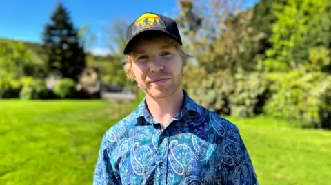 Ash Carter is standing in a green field on a sunny day with bushes and trees behind. He has sandy blonde hair and stubble. He is wearing a black cap with a yellow and orange logo on the front and he is wearing a blue shirt with white patterns.
