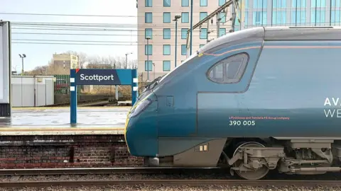 AvantiWestCoast A blue sign for Stockport station, re-written to say Scottport in white lettering, erected outdoors on a train station platform, with a train car seen on railway tracks in the foreground. 