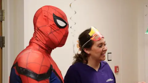 Cambridge University Hospitals Spiderman with play specialist Sophie Barber on the hospital ward. Sophie wears purple scrubs and has a wonder woman mask pulled onto her face. She has brown hair which has been pulled away from her face by a claw clip.