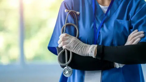 Getty Images A female doctor, pictured from the neck down, in blue scrubs with stethoscope around her neck