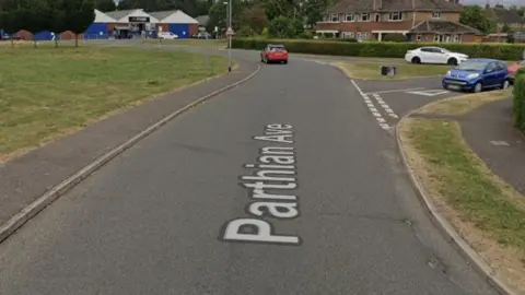 Google A Google street view image of a road with a grass area to the left and houses and cars to the right.