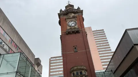 An image of the brick tower with a clock at the top against a grey sky with grey buildings either side. A high rise block of flats is visible in the background
