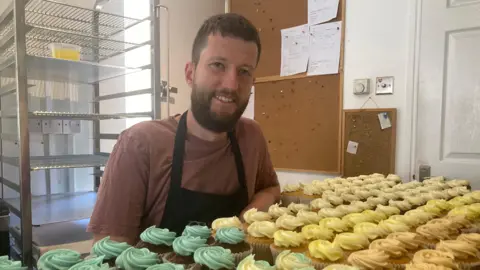 A man with a beard stand behind a large tray of cupcakes, decorated with green and yellow icing.