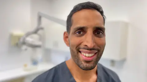 Mark Norman BBC A dentist wearing dental scrubs smiling at the camera with dental equipment in the background