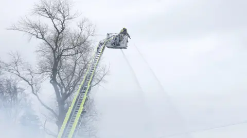 Pacemaker Press. Un bombero está en lo alto de una escalera aérea vertiendo agua con una manguera sobre un incendio. Al fondo se ve un árbol sin hojas.