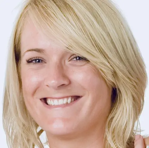Family A woman with blonde hair smiles at the camera in front of a white background; the picture shows her head and shoulders