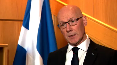 John Swinney standing beside a Saltire, he is bald, has glasses and a dark suit and tie.